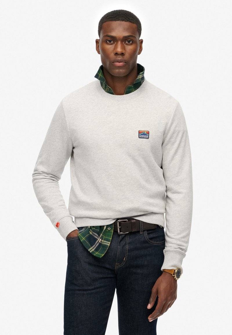 Sudadera gris con cuello redondo, superpuesta a una camisa de cuadros verde. Notable parche de logo en el pecho, con jeans oscuros y un cinturón negro.