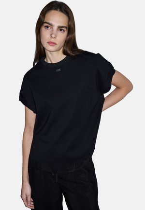 Jonge vrouw met lang bruin haar, gekleed in een loszittend zwart shirt met korte mouwen en een zwarte broek, staand met één hand op haar heup.