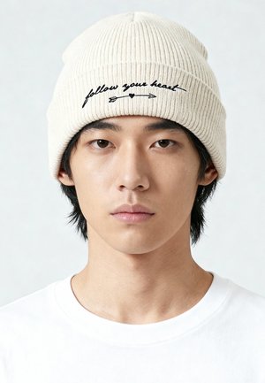 Roomkleurige geribbelde muts met geborduurde tekst "volg je hart" en pijlontwerp, gedragen boven een wit shirt, tegen een lichte achtergrond.