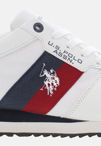 Sneaker bianco con strisce rosse e blu navy, dotata di un logo polo argentato, tessuto testurizzato e occhielli rinforzati. Inclusa la brandizzazione U.S. Polo Assn.