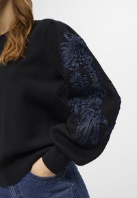 Sweatshirt noir avec des détails en broderie texturée bleu marine sur la manche gauche et des poignets côtelés, associé à un jean en denim bleu.