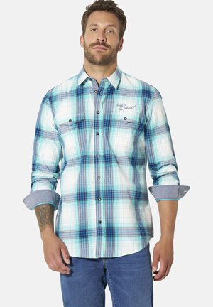 Mann trägt blau-weiß kariertes Hemd mit aufgekrempelten Ärmeln und blaue Jeans, steht vor einfachem Hintergrund.
