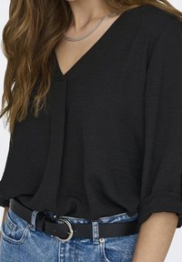 Black, loose-fitting blouse with a V-neckline. Made of textured fabric, featuring rolled sleeves. Paired with high-waisted denim and a black belt.
