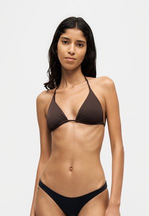 Femme aux longs cheveux foncés portant un haut de bikini marron foncé et un bas de bikini noir sur un fond clair uni.