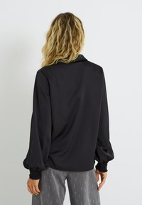 Blusa negra ligera con textura suave, que cuenta con cuello y puños fruncidos. La parte trasera tiene un ajuste relajado con un ligero drapeado.