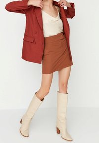 Femme ajustant un blazer couleur rouille sur un haut drapé beige, portant une mini-jupe assortie couleur rouille et des bottes crème montantes jusqu'aux genoux avec talons carrés.