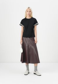 Young woman wearing black embellished sleeve t-shirt, brown leather midi skirt, gray socks, white loafers, holding small white handbag.