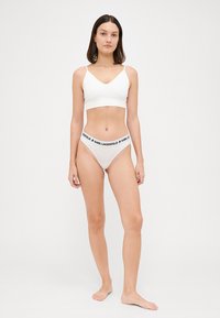 Biely základný bralet s tenkými ramenami a zodpovedajúcim bielym tangami s čiernym elastickým pásom s logom. Hladký, ľahký materiál.