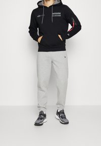 Felpa nera con fodera interna grigia, tasca frontale, logo dei Buccaneers e tasca laterale con zip. Abbinata a pantaloni della tuta grigi e scarpe da ginnastica nere.