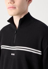 Μαύρο, υφαντό φούτερ με φερμουάρ που διαθέτει ψηλό γιακά, λευκές ρίγες και ένα μικρό λογότυπο "BOSS" στο στήθος.