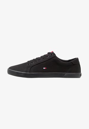 Sneakers neri in tela con texture liscia, punta rotonda e suola piatta in gomma. Presentano occhielli metallici e un piccolo patch con logo sul lato.