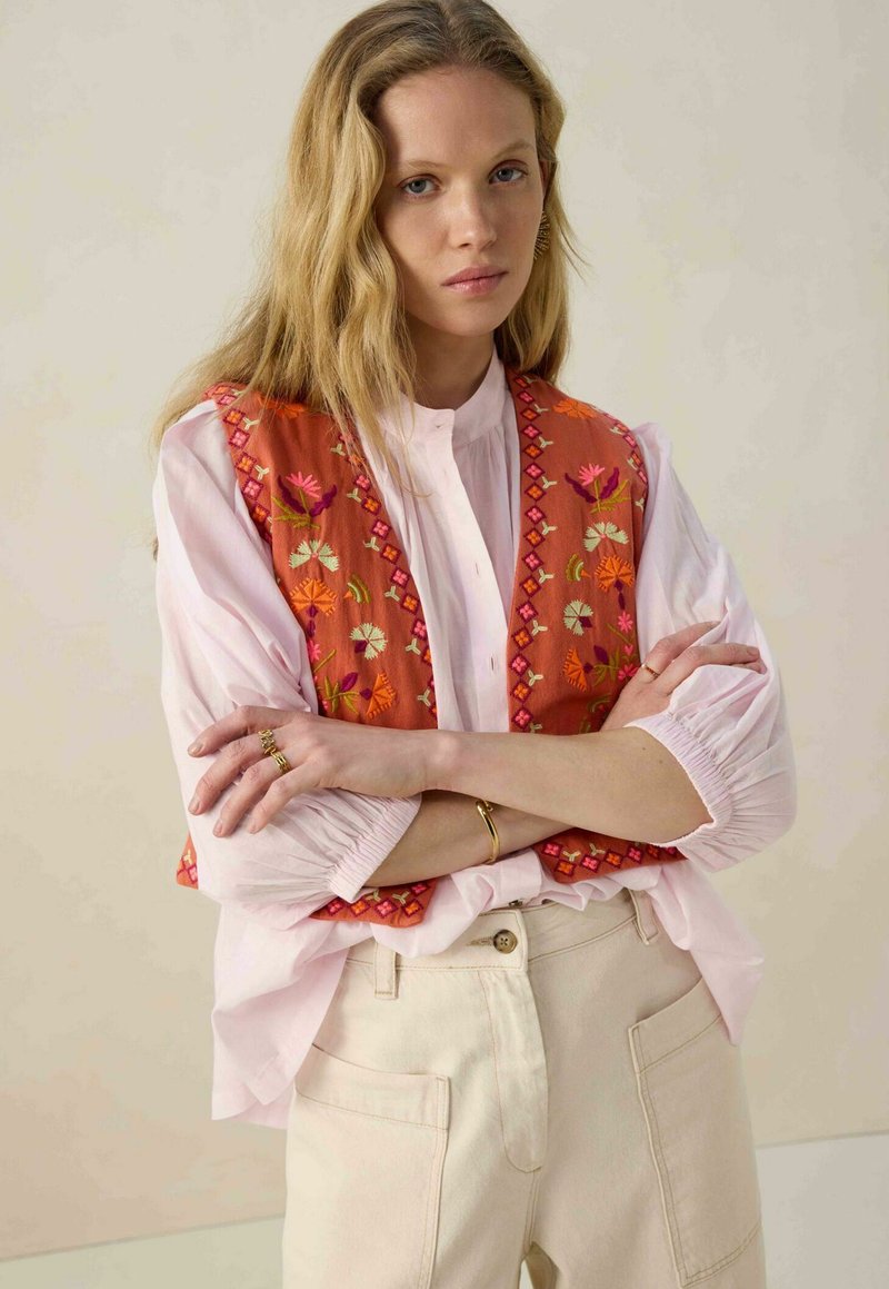 Gilet orange brodé avec des motifs floraux sur une blouse rose clair. La tenue présente des manches bouffantes et un pantalon large de couleur crème.