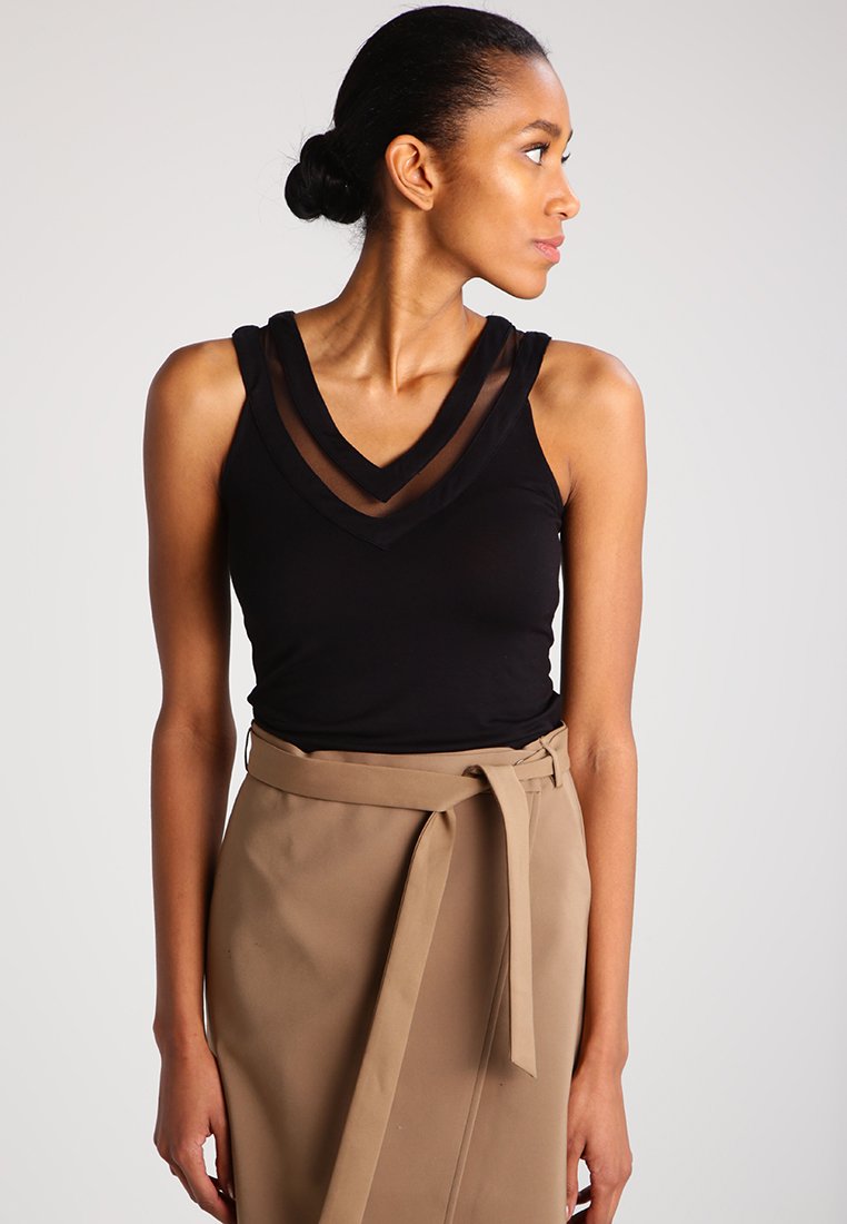 Haut noir sans manches avec décolleté en V, présentant des accents transparents noirs. Porté avec une jupe portefeuille beige, soulignant des lignes épurées et un design moderne.