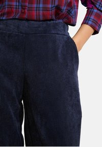 Jūras spēku krāsas corduroy bikses ar teksturētu apdari, aprīkotas ar elastīgu jostasvietu un sānu kabatām, kas saskaņotas ar raibs kreklu virsū.