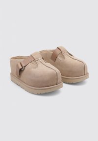 Zoccoli in suede beige con punta arrotondata, dotati di una fascia in pelle marrone e una suola in gomma testurizzata per una migliore aderenza.