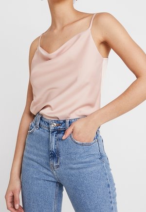 Femme portant un caraco en satin rose clair à fines bretelles, rentré dans un jean en denim bleu taille haute, main dans la poche.