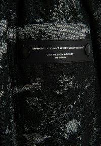 Tejido con un patrón en blanco y negro con un aspecto texturizado, que presenta una etiqueta negra con texto blanco y botones. Visible branding de la colaboración de diseño.