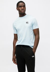 Gaiši zils īsajām piedurknēm T krekls ar apaļu kakla izgriezumu un mazu melnbaltu logo uz krūšu daļas, apvienots ar melniem biksēm ar saistošu jostu.