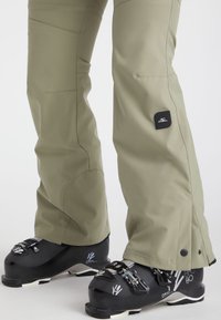 Beige skibroek met een taps toelopend ontwerp en zijzakken, gecombineerd met zwarte skischaatsen met verstelbare gespen en witte graphics.