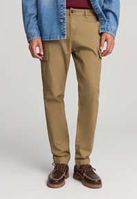 Pantaloni cargo color khaki con tasche laterali, vestibilità affusolata e tessuto in cotone, abbinati a una camicia di denim e scarpe marroni con design alla allacciatura.