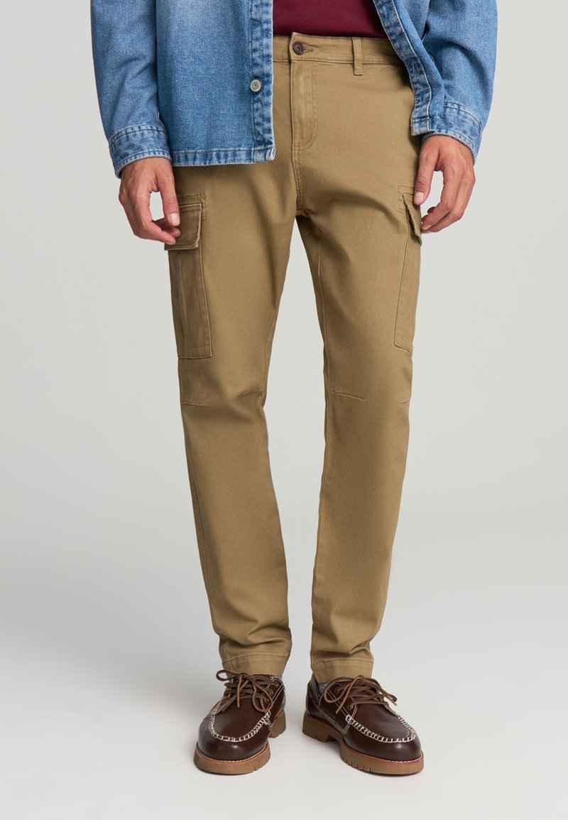 Pantaloni cargo color khaki con tasche laterali, vestibilità affusolata e tessuto in cotone, abbinati a una camicia di denim e scarpe marroni con design alla allacciatura.