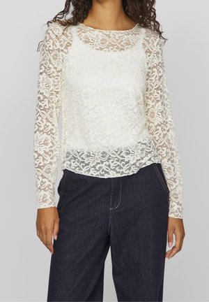Femme portant un chemisier en dentelle florale blanche à manches longues et un jean sombre taille haute, debout devant un fond uni.