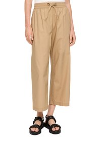 Beige wijde broek met een elastische tailleband en koord, gemaakt van een lichtgewicht stof. Gecombineerd met zwarte gevlochten sandalen.