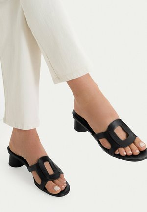 Stopy w czarnych sandałach na obcasie z odkrytymi palcami, szerokimi paskami i geometrycznymi wycięciami, zestawione z białymi krótkimi spodniami.