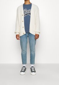 Rödpullover i chunky vitstickad med V-ringning, med stora knappar; blå t-shirt med grafiskt tryck under; ljusbl å tapered jeans; sneakers.