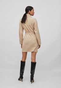 Robe en tricot beige avec une taille ceinturée, des manches longues et un col haut. Portée avec des bottes noires brillantes jusqu'aux genoux. Texture douce, design minimal.