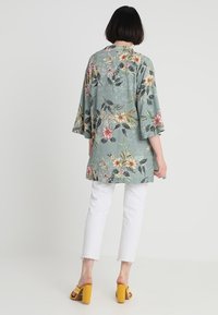 Floral kimono jacket in light green fabric, featuring large multicolored flowers and leaves. Worn over white cropped pants and yellow heels.