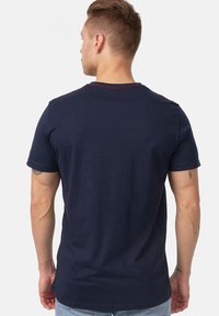 Mørkeblå bomulds t-shirt med korte ærmer. Har en rund hals med rød syning og en glat tekstur. Synlige tatoveringer på venstre arm.
