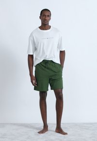 Πράσινα σορτς με κορδόνι στη μέση και λευκό μπλουζάκι με το λογότυπο "TOMMY HILFIGER". Το μοντέλο στέκεται ξυπόλυτο σε ανοιχτό γκρι χαλί.