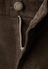 Bruine corduroy stof met een zichtbare metalen rits en een knop met de inscriptie 'opgericht in 1984', die textuur en stikdetails benadrukt.