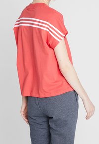 Camiseta de algodón color coral con mangas cortas, que presenta rayas blancas en la parte posterior. Combinada con pantalones grises, que muestran un diseño y un corte relajado.
