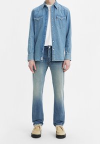 Hombre vistiendo una camisa de denim azul claro sobre una camiseta blanca, con jeans azul desteñido y zapatos beige de cordones, frente a un fondo blanco.