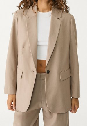 Vrouw met een beige, op maat gemaakte blazer en bijpassende broek, een wit cropped geribd topje, staande met ontspannen handen langs haar zij.