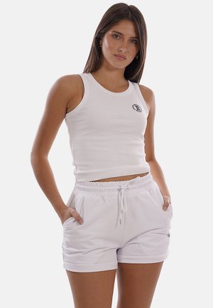 Giovane donna con capelli lisci che indossa un top corto bianco senza maniche e pantaloncini bianchi con coulisse, con le mani nelle tasche.