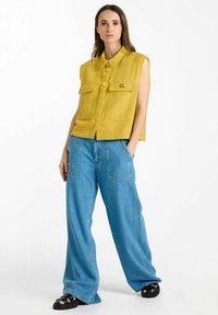 Haut sans manches jaune à boutons avec deux poches avant, associé à un pantalon en denim bleu large et des sandales noires sur un fond uni.