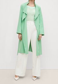 Długi płaszcz w kolorze mięty z kołnierzem z marszczeniem, zestawiony z jasnzie zieloną bluzką i białymi szerokimi spodniami. Buty są kremowe, na spiczastym nosku.