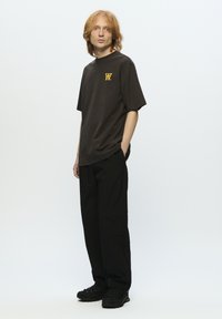 Dunkelbraunes Baumwoll-T-Shirt mit kurzen Ärmeln und gelbem Logo auf der Brust, kombiniert mit schwarzen, locker sitzenden Hosen und schwarzen Sneakers.