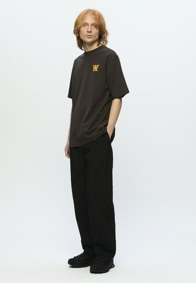 T-shirt a maniche corte in cotone marrone scuro con un logo giallo sul petto, abbinato a pantaloni larghi neri e sneakers nere.