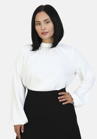 Hvid langærmet top med rynket halsudskæring og flagrende ærmer, parret med en sort højlivskørt. Subtil glans tekstur synlig.