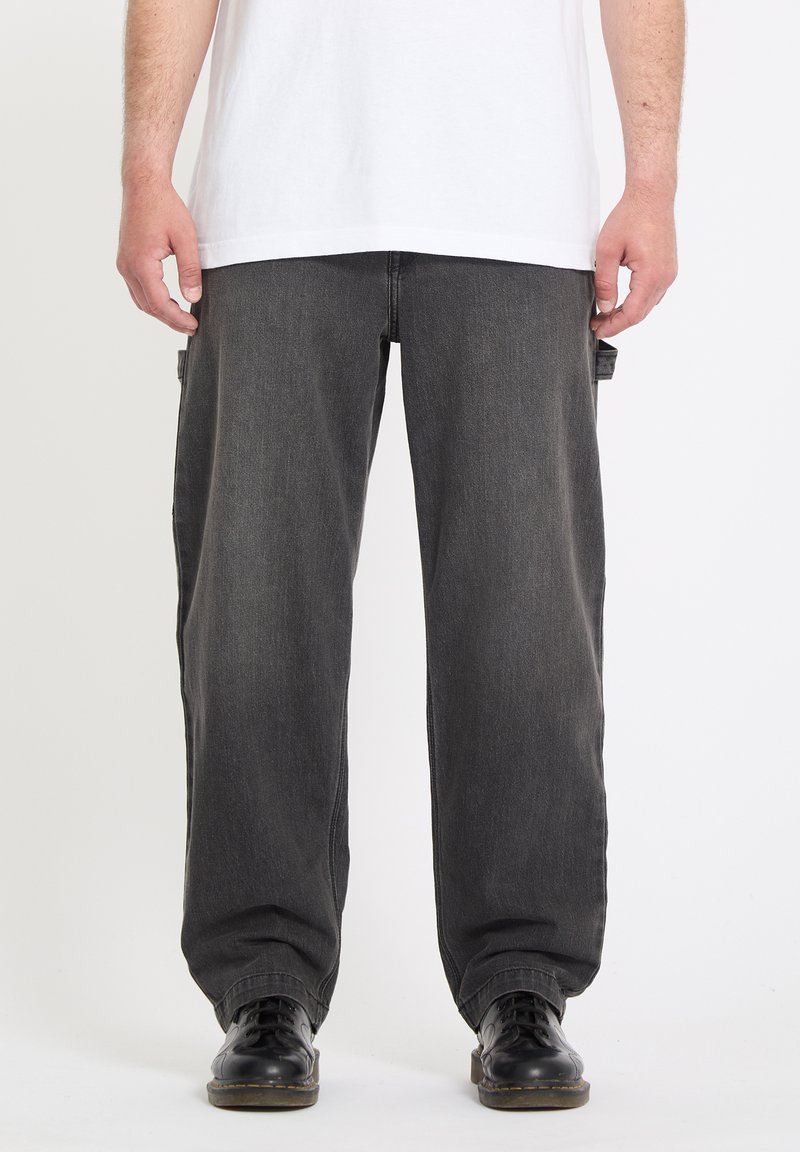 Homme portant un jean noir ample, des chaussures noires à lacets et un T-shirt blanc uni, debout devant un fond blanc.