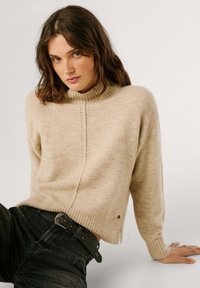 Ljusbeige stickad tröja med hög krage, ribbat avslut och manschetter, med en vertikal sömdetalj. Bärs över mörka jeans med ett svart bälte.