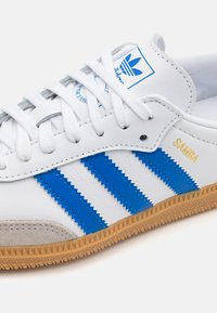 Hvit lær sneaker med blå striper og suedes tåhette, med flate hvite snørebånd og teksturert gummisål. "Samba"-logo i gull.
