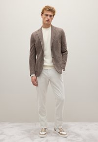 Braun kariertes Blazer, weißer Pullover, hellgraue gerippte Hose und beige Sneakers mit weißen Sohlen und goldenen Akzenten, vor einem schlichten Hintergrund stehend.