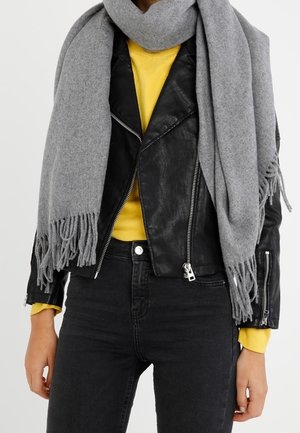 Personne portant une écharpe grise à franges sur une veste en cuir noire avec des fermetures éclair argentées, un t-shirt jaune à manches longues et un jean noir.