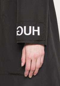 Giacca nera con grande logo bianco "HUGO" sul polsino, dalla texture liscia e opaca e vestibilità sartoriale.