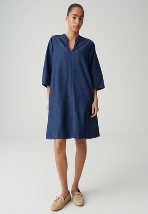 Femme debout portant une robe en jean bleu foncé ample jusqu'aux genoux avec des manches trois-quarts et des mocassins beiges.